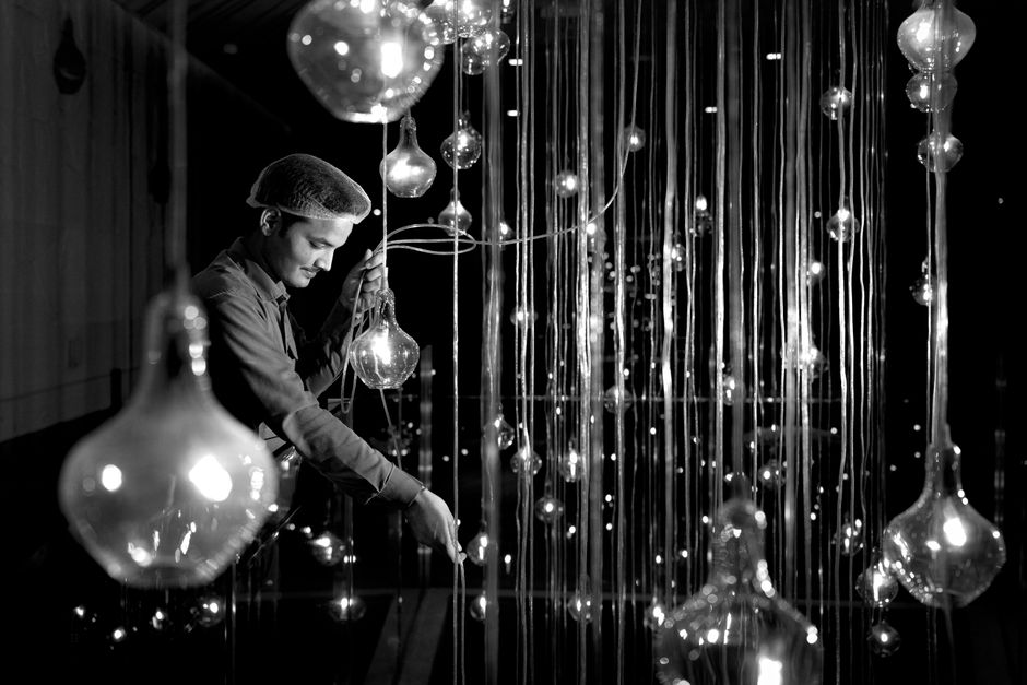 A man adjusts an intricate light bulb display, with reflections creating a stunning visual effect.