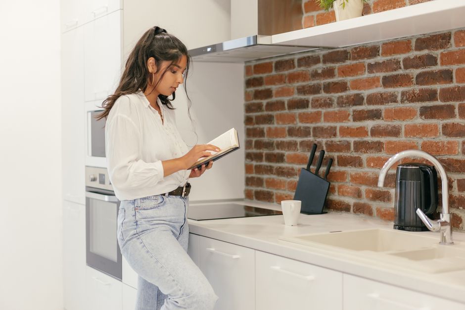A woman reads a book while leaning against a countertop in a modern kitchen.